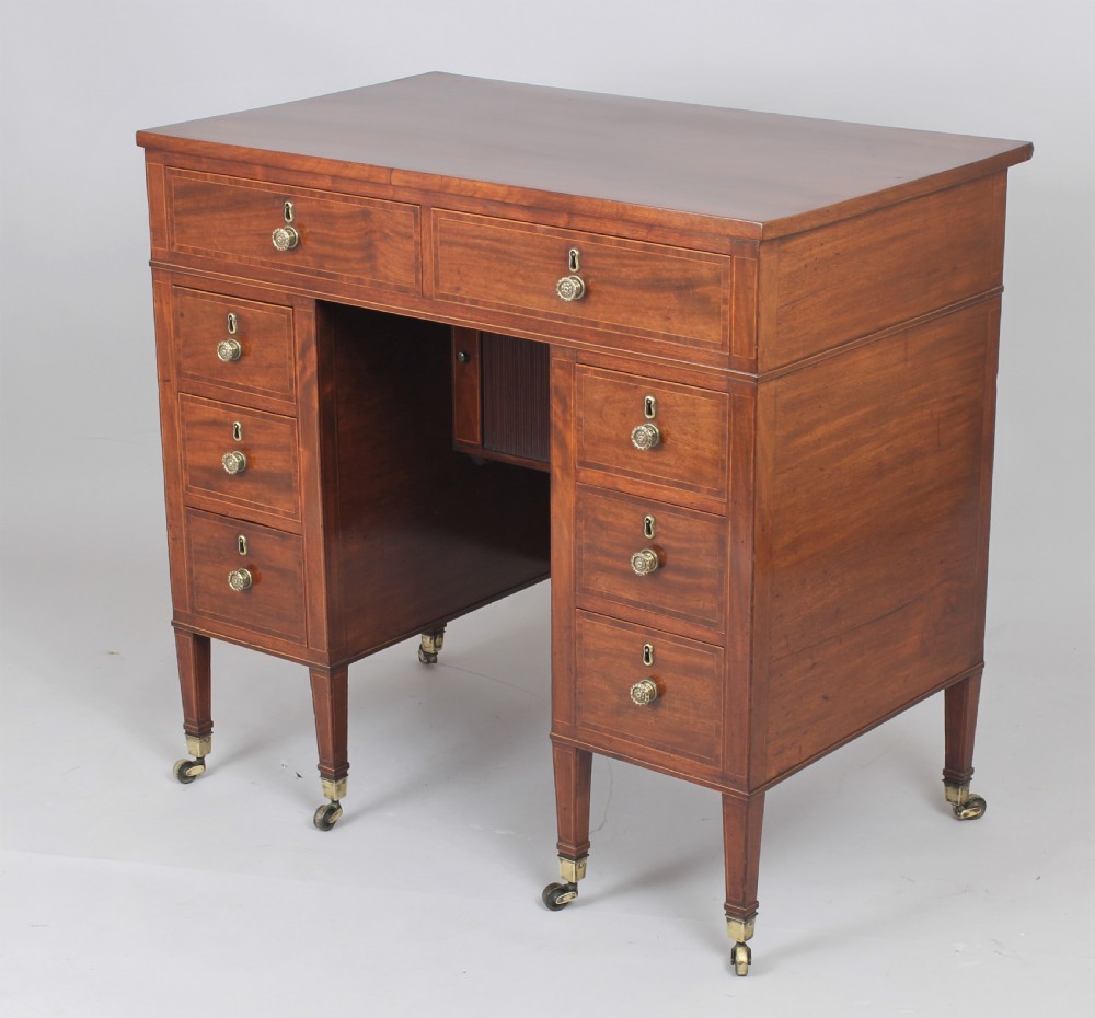 A late 18th-century Sheraton period mahogany kneehole desk featuring multiple drawers with brass handles, inlaid boxwood stringing, and square tapered legs on castors.
