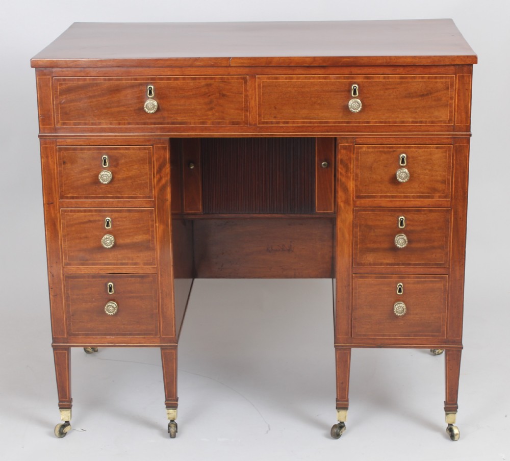 A late 18th-century Sheraton period mahogany kneehole desk featuring multiple drawers with brass handles, inlaid boxwood stringing, and square tapered legs on castors. View II