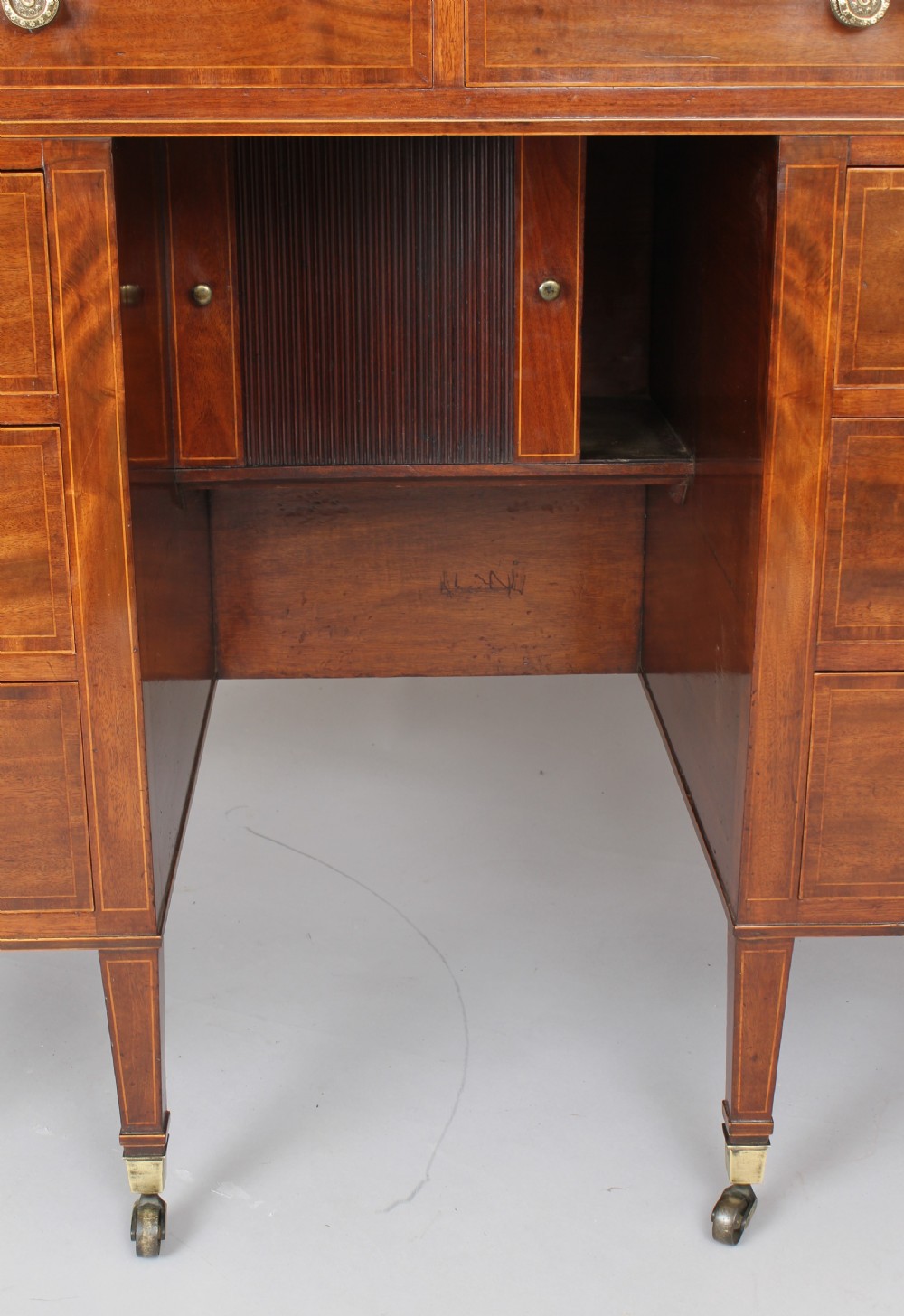 A late 18th-century Sheraton period mahogany kneehole desk featuring multiple drawers with brass handles, inlaid boxwood stringing, and square tapered legs on castors. View III