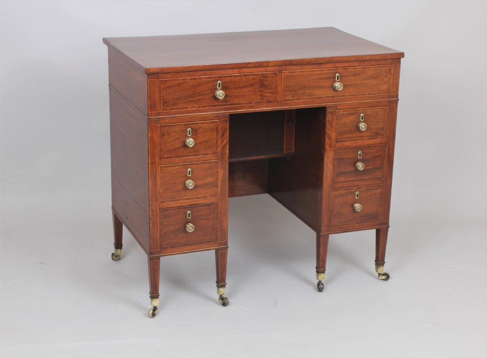 A late 18th-century Sheraton period mahogany kneehole desk featuring multiple drawers with brass handles, inlaid boxwood stringing, and square tapered legs on castors. View VI