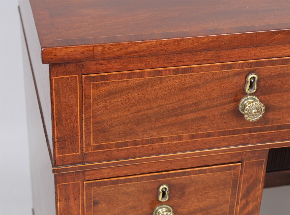 A late 18th-century Sheraton period mahogany kneehole desk featuring multiple drawers with brass handles, inlaid boxwood stringing, and square tapered legs on castors. View VII