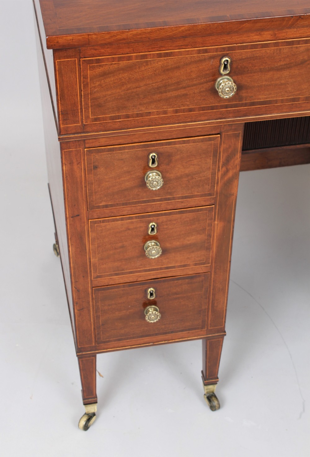 A late 18th-century Sheraton period mahogany kneehole desk featuring multiple drawers with brass handles, inlaid boxwood stringing, and square tapered legs on castors. View VIII