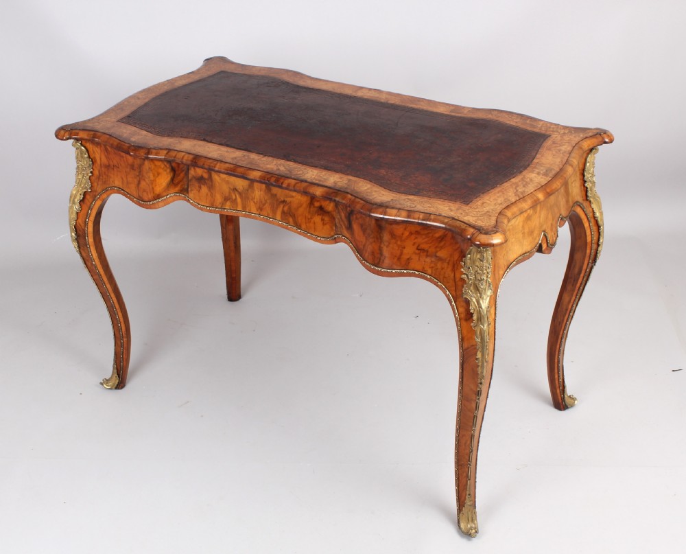 A French Rococo style burr walnut writing table featuring cabriole legs, ornate brass mounts, and a dark brown leather writing top.