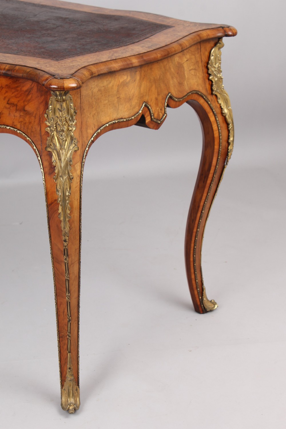 A French Rococo style burr walnut writing table featuring cabriole legs, ornate brass mounts, and a dark brown leather writing top. View III