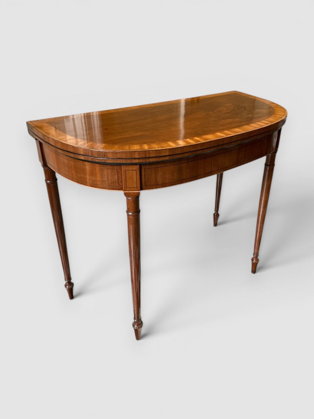 An early 19th-century mahogany demilune card table featuring satinwood crossbanding and turned tapered legs.