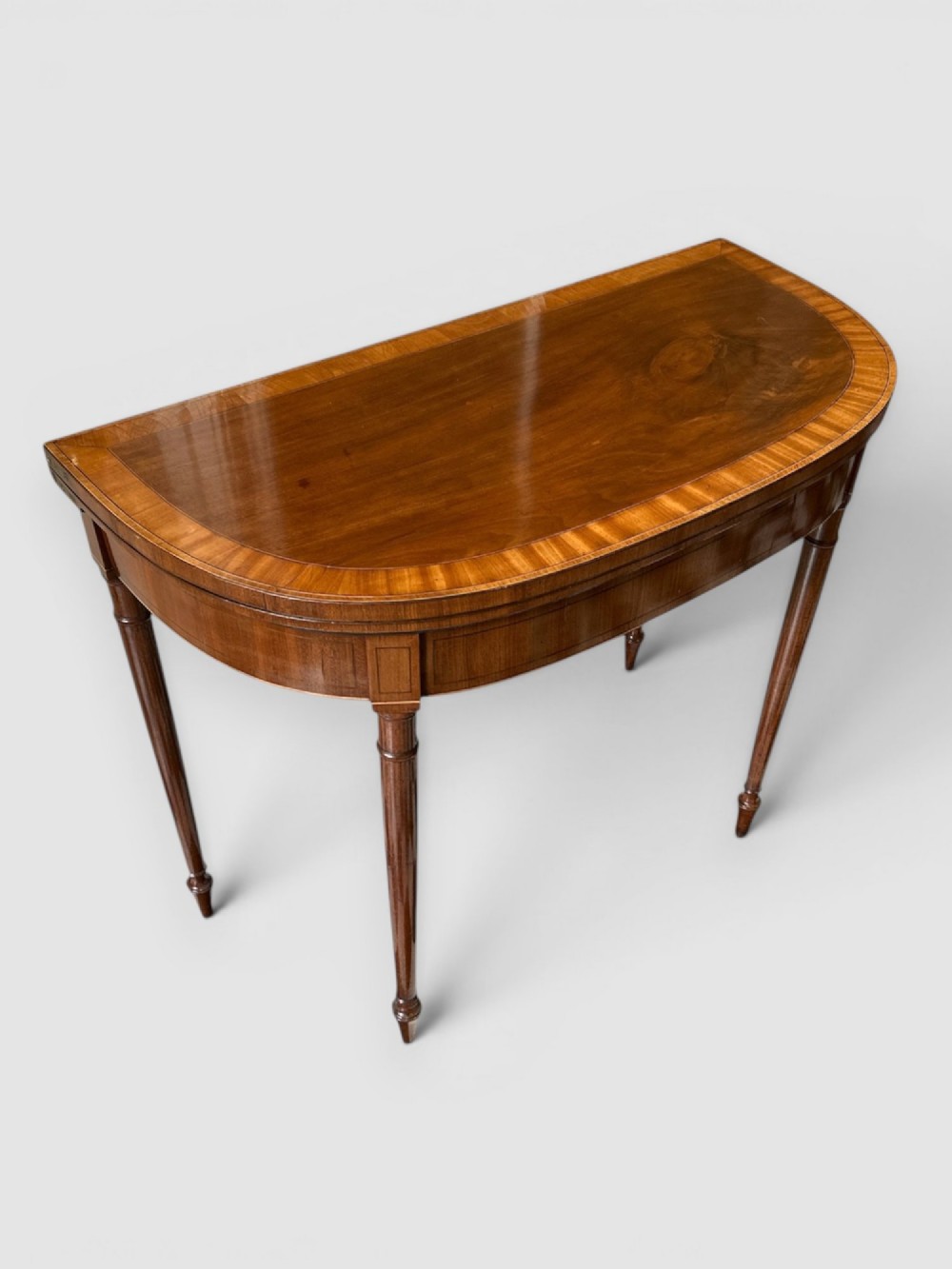 An early 19th-century mahogany demilune card table featuring satinwood crossbanding and turned tapered legs. View II