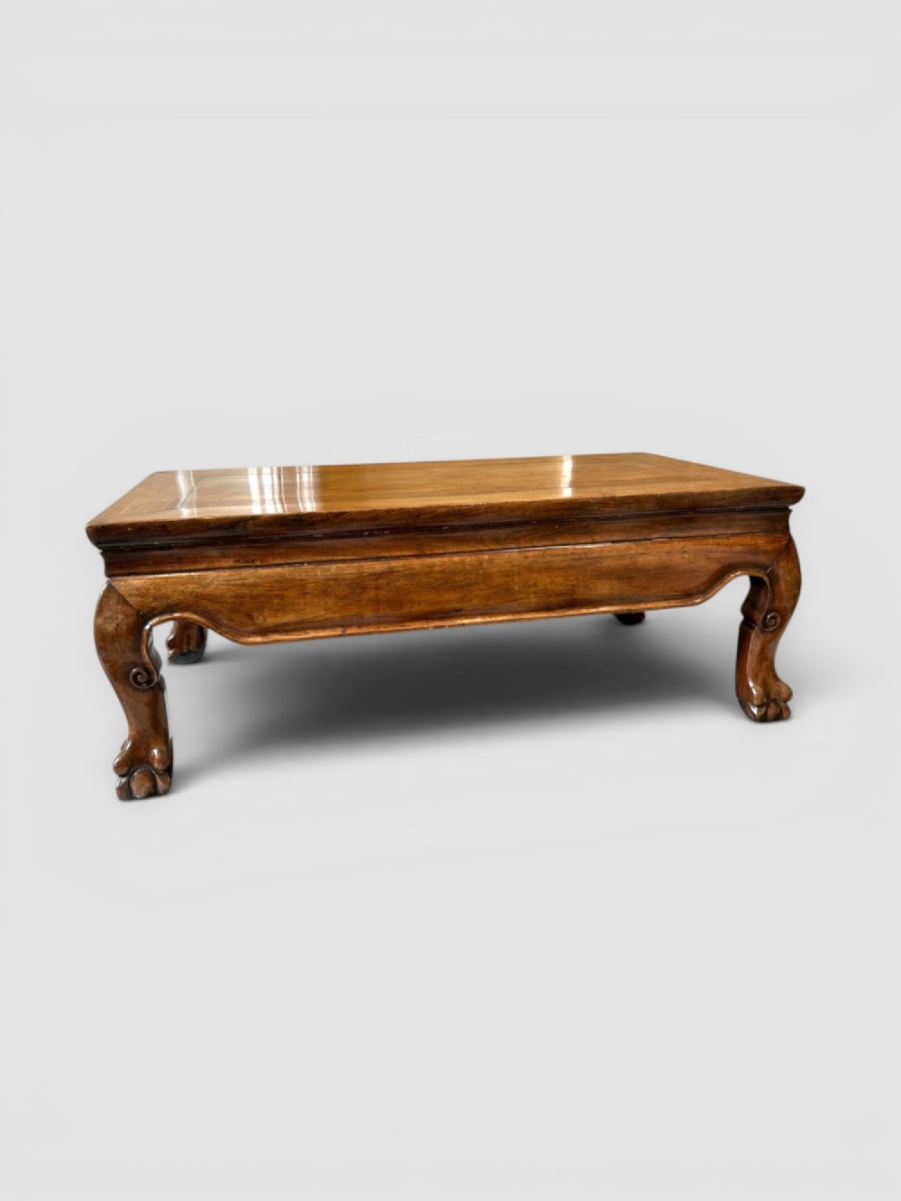 A low rectangular warm brown wooden Chinese opium table featuring short cabriole legs, carved scrolled feet, and a polished panel top.