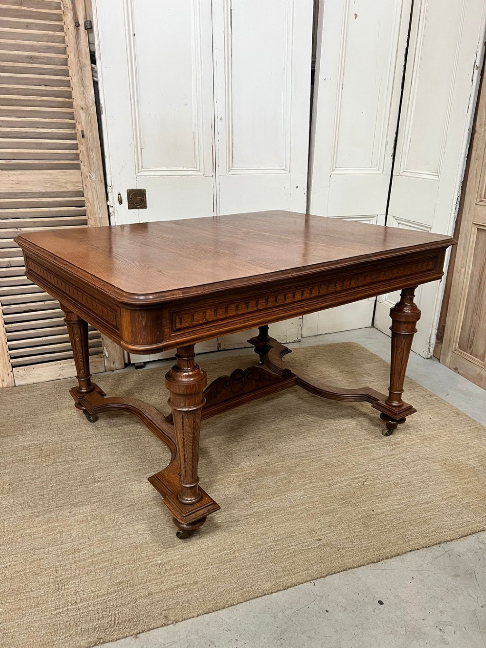 An antique French fruitwood dining table featuring turned legs, a carved apron, a low stretcher base, and small metal castors.