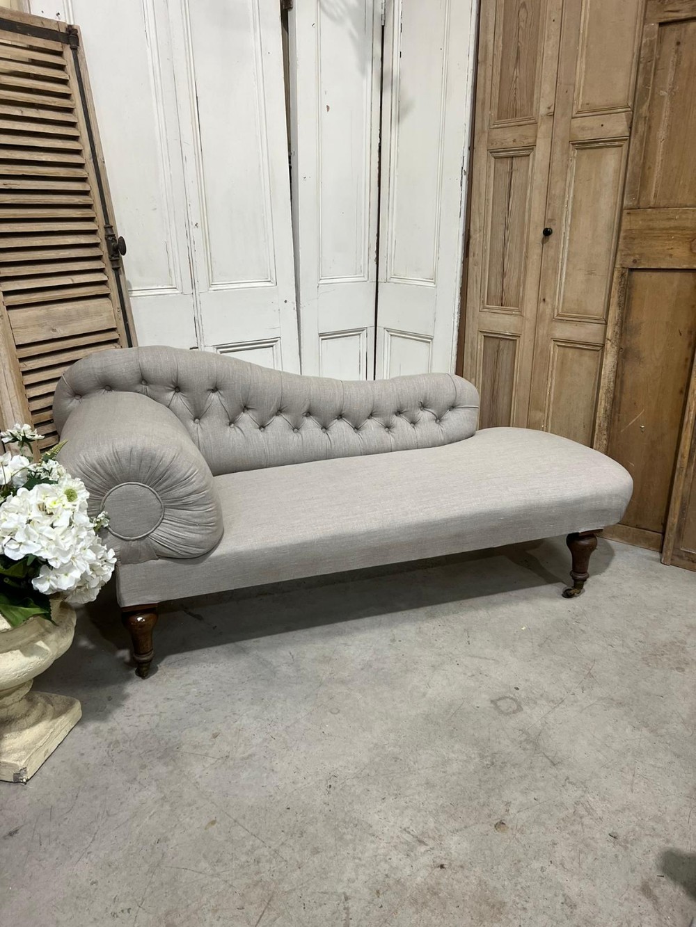 A Victorian chaise longue upholstered in neutral grey linen fabric featuring deep buttoning along the contoured backrest, a scroll arm, turned dark wood legs, and brass castors.