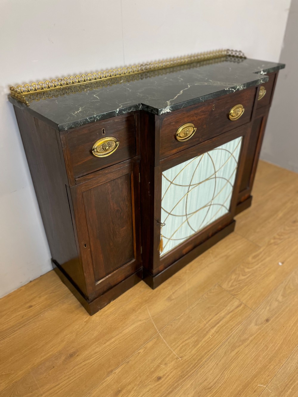 An early nineteenth-century Regency rosewood secretaire side cabinet featuring a dark green marble top with a brass gallery, a pleated fabric door panel, and brass handles.