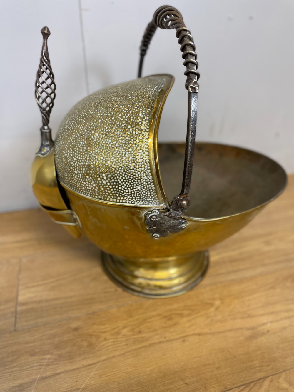 An Arts and Crafts style brass coal helmet with a textured hood, a wire-wrapped swing handle, a round pedestal base, and a matching hand shovel. View V