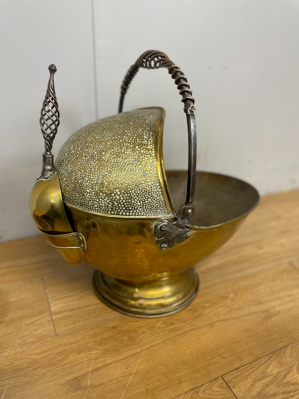 An Arts and Crafts style brass coal helmet with a textured hood, a wire-wrapped swing handle, a round pedestal base, and a matching hand shovel. View X