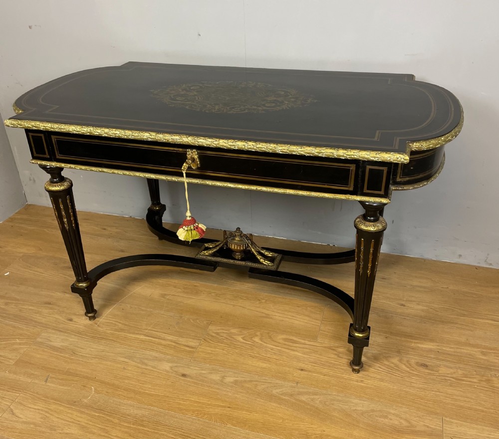 An ebonised nineteenth-century French writing table featuring gilt-bronze mounts, fluted tapered legs, a single central drawer, and a shaped top with central marquetry detailing.