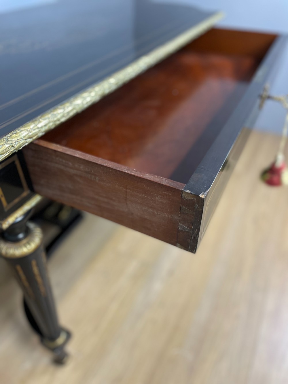 An ebonised nineteenth-century French writing table featuring gilt-bronze mounts, fluted tapered legs, a single central drawer, and a shaped top with central marquetry detailing. View IX