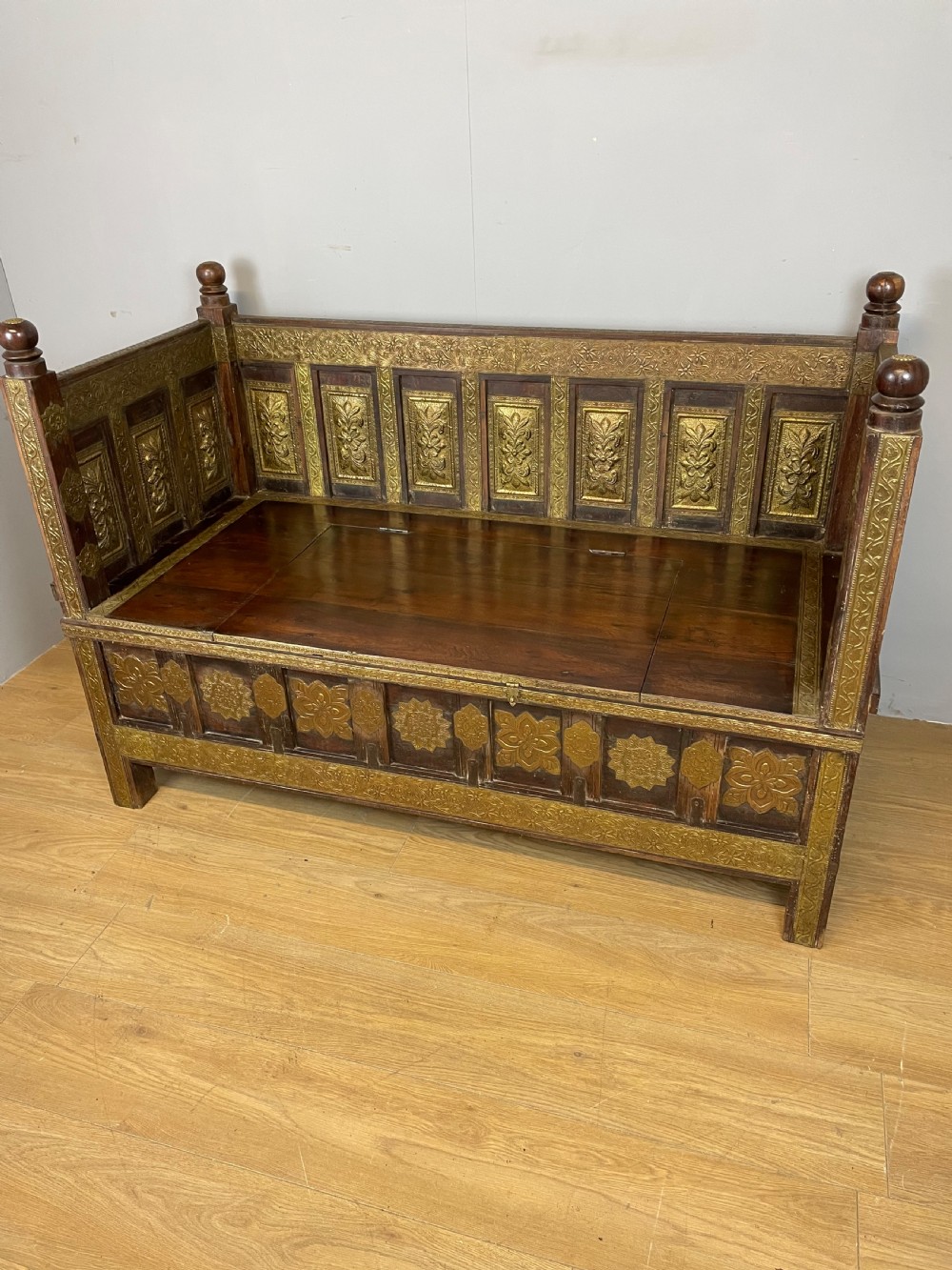 An Anglo Indian dark hardwood box settle featuring embossed brass panels, turned corner finials, and a hinged lift-top seat.