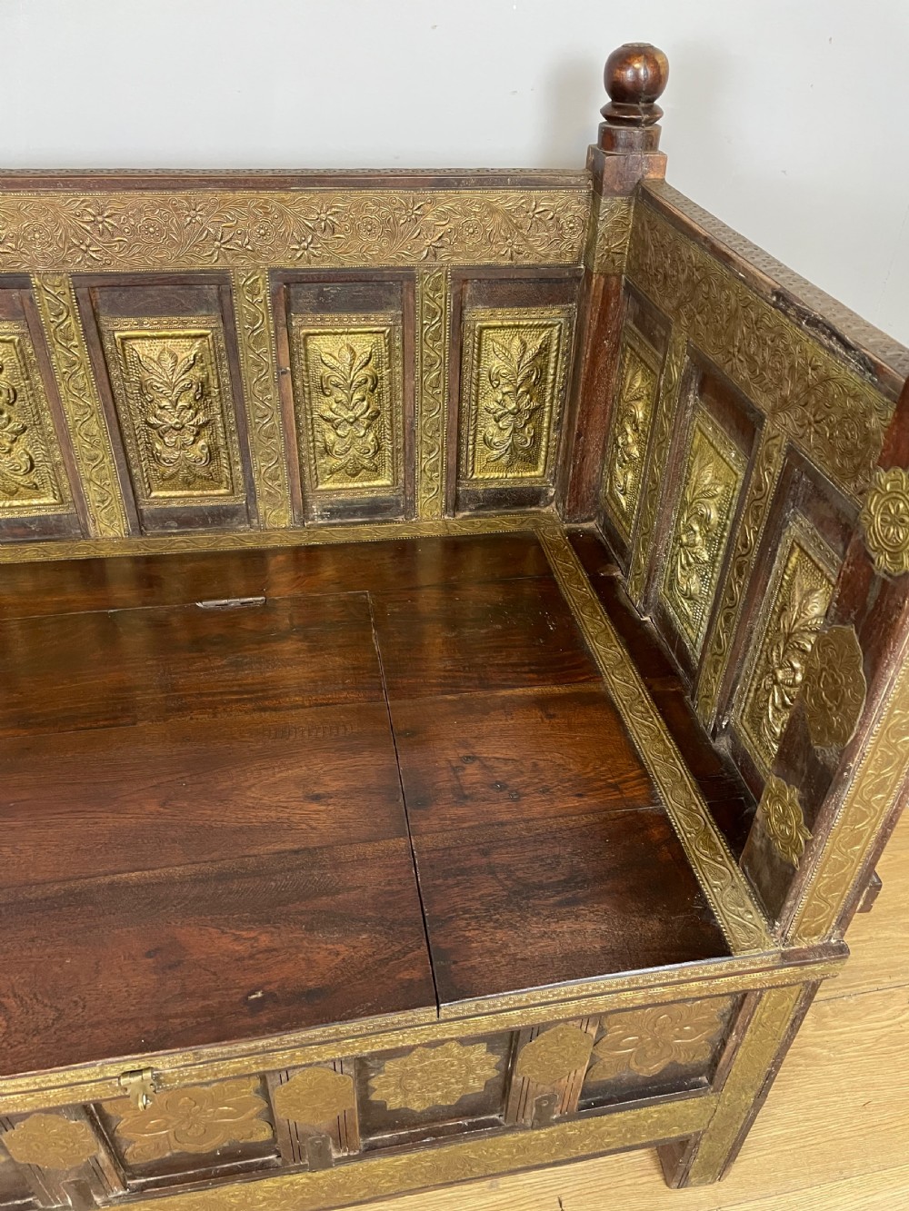 An Anglo Indian dark hardwood box settle featuring embossed brass panels, turned corner finials, and a hinged lift-top seat. View II
