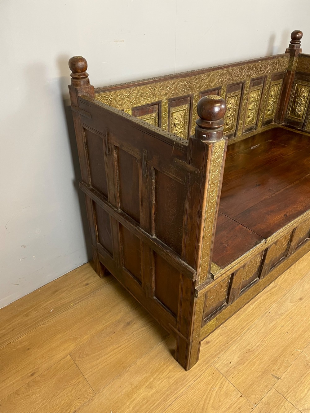 An Anglo Indian dark hardwood box settle featuring embossed brass panels, turned corner finials, and a hinged lift-top seat. View V