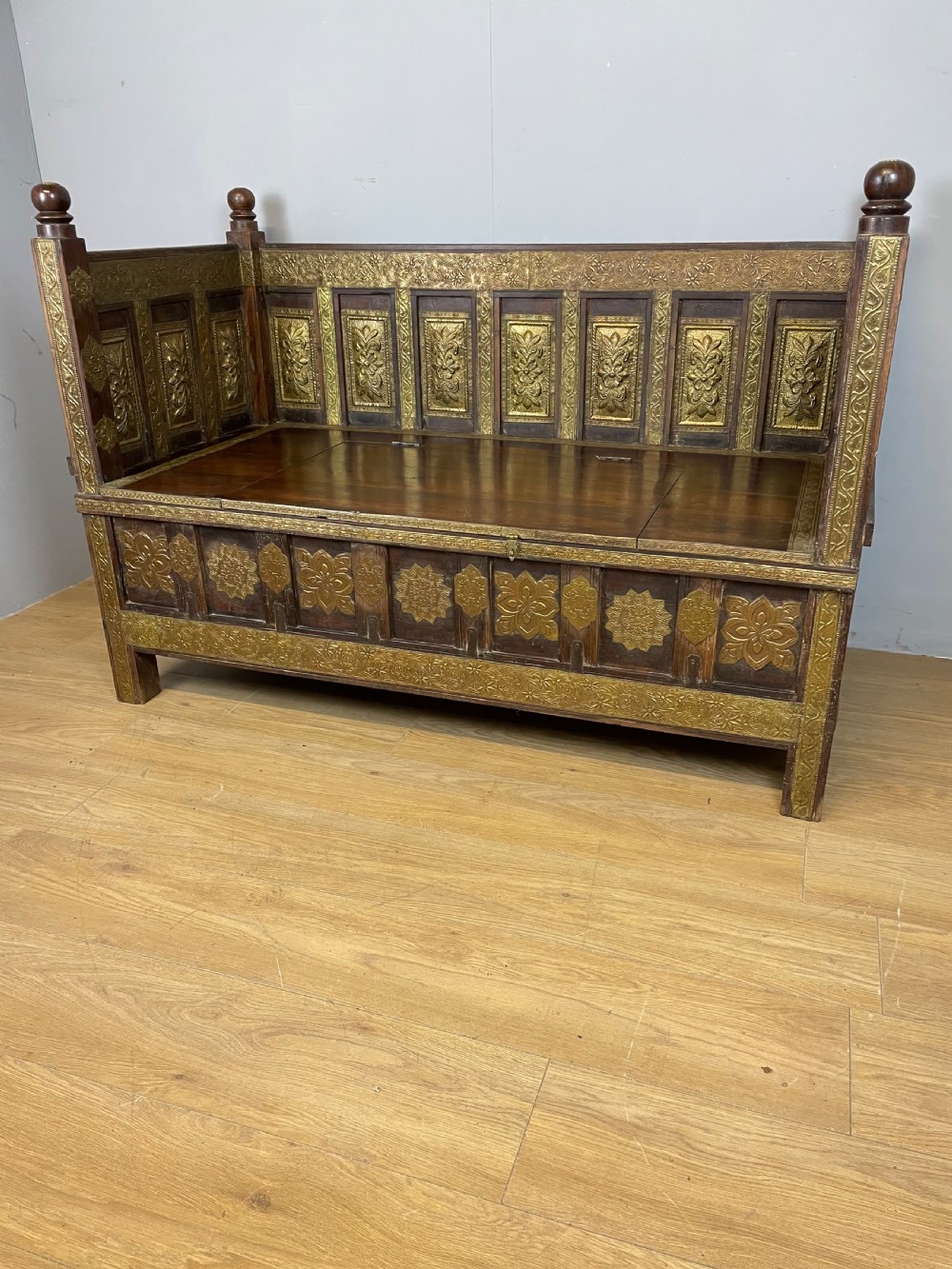 An Anglo Indian dark hardwood box settle featuring embossed brass panels, turned corner finials, and a hinged lift-top seat. View VI