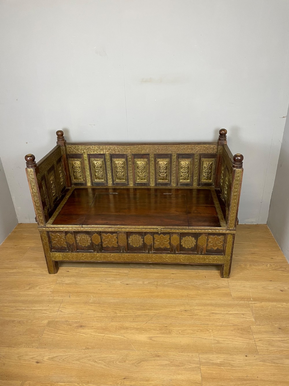 An Anglo Indian dark hardwood box settle featuring embossed brass panels, turned corner finials, and a hinged lift-top seat. View XIII