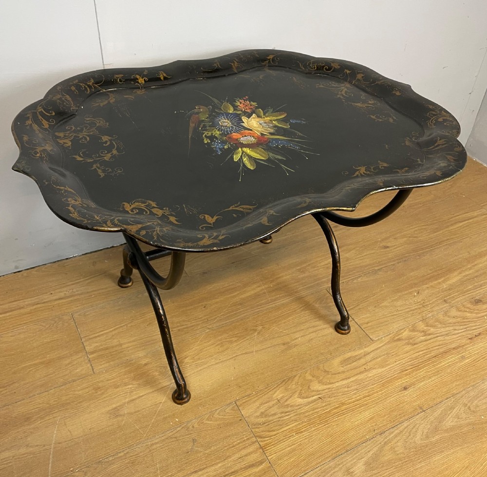 A nineteenth-century black lacquered papier-mache tray with scalloped edges and a central hand-painted floral motif resting on a black iron stand.
