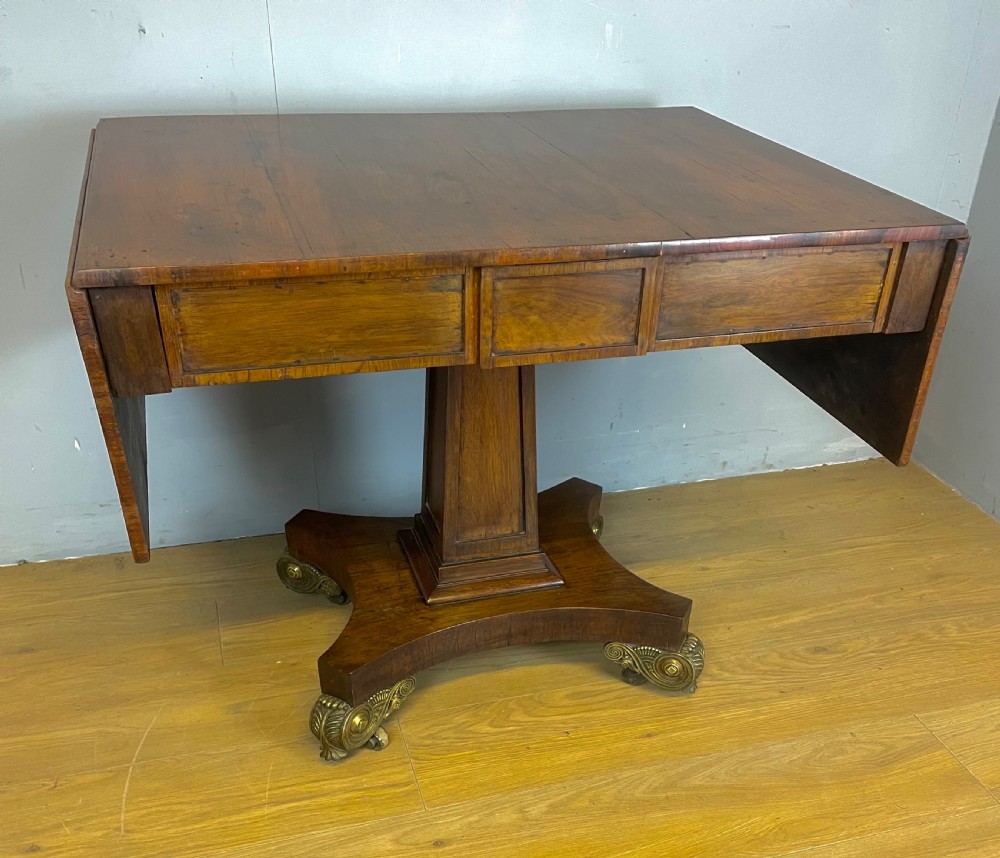 A Regency rosewood drop-leaf sofa table featuring two frieze drawers, a central column, and a quadruped platform base with decorative brass castors.