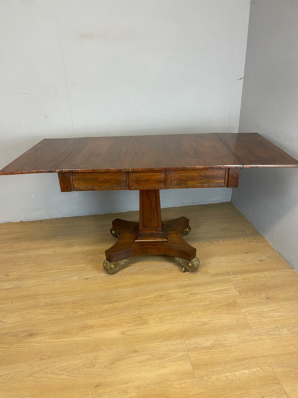 A Regency rosewood drop-leaf sofa table featuring two frieze drawers, a central column, and a quadruped platform base with decorative brass castors. View XI
