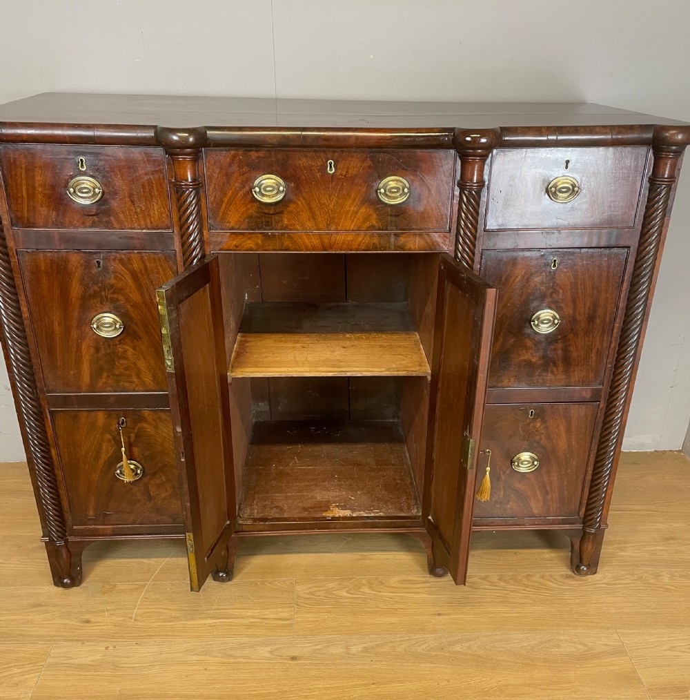 A Regency flame mahogany sideboard featuring rope-twist corner columns, multiple drawers with brass ring handles, and central cupboard doors. View VI