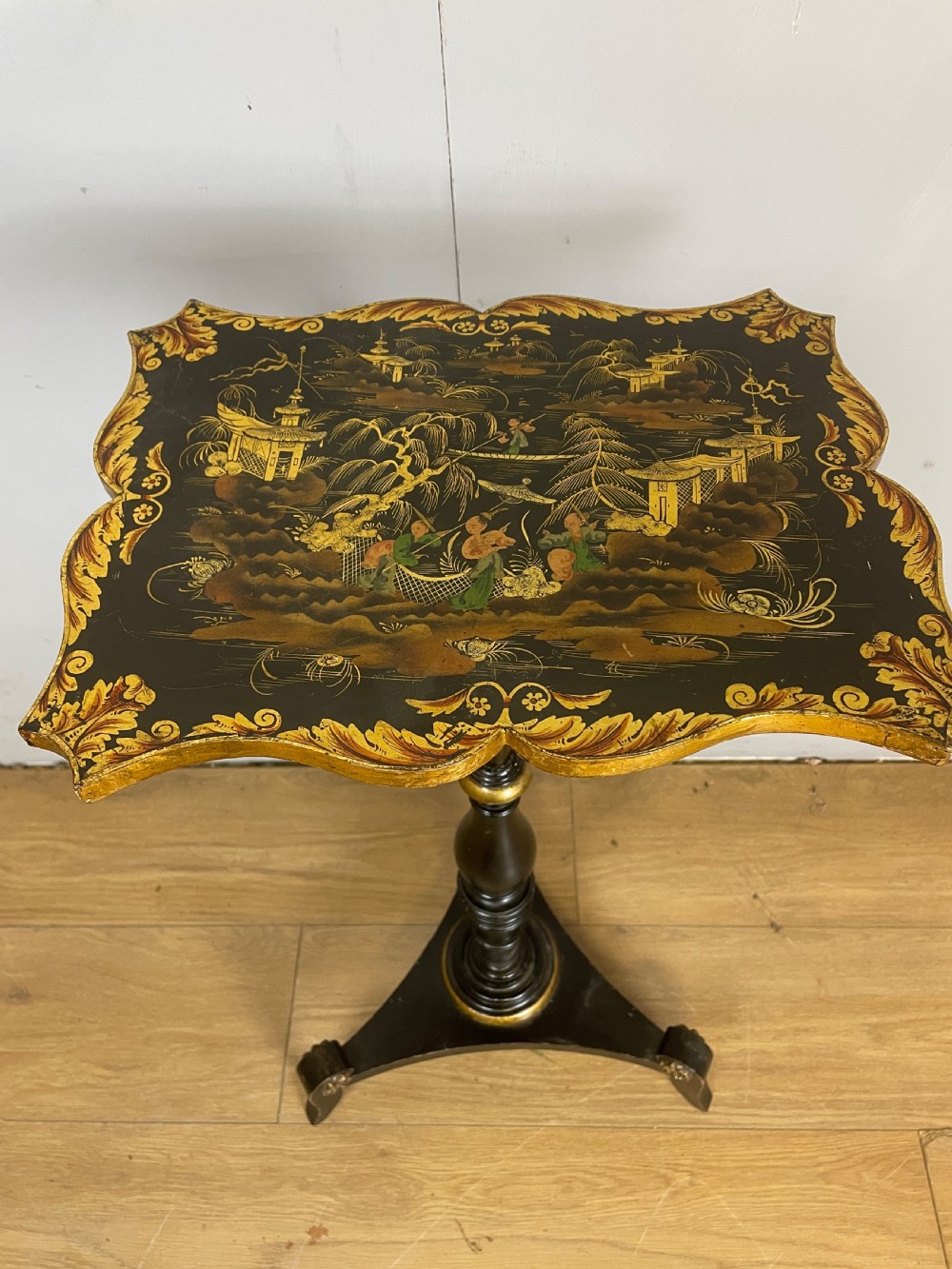 A nineteenth-century Chinoiserie wine table featuring a scalloped black lacquered top with gilded oriental landscape decoration, supported by a turned ebonised column on a tripartite base. View II