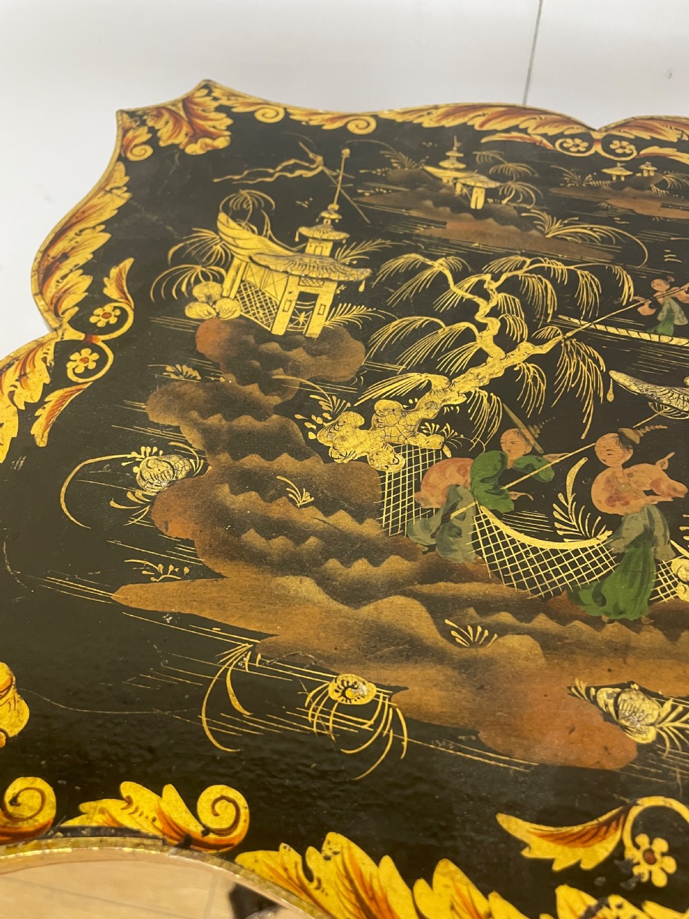 A nineteenth-century Chinoiserie wine table featuring a scalloped black lacquered top with gilded oriental landscape decoration, supported by a turned ebonised column on a tripartite base. View X