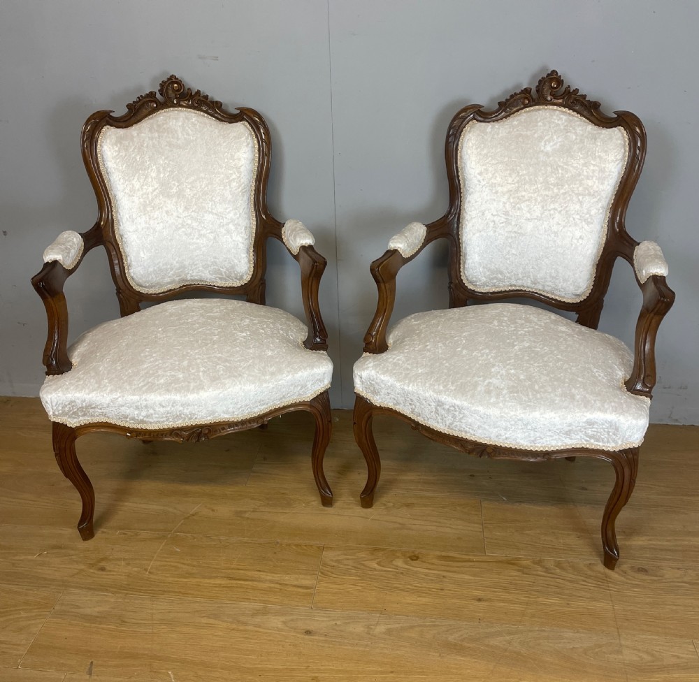 A pair of French carved walnut open armchairs upholstered in cream crushed fabric with cabriole legs and ornate cresting.