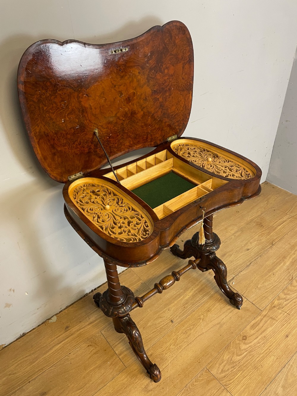A Victorian burr walnut work table shown with its hinged lid open, revealing intricate fretwork covers, internal compartments, and carved turned legs.