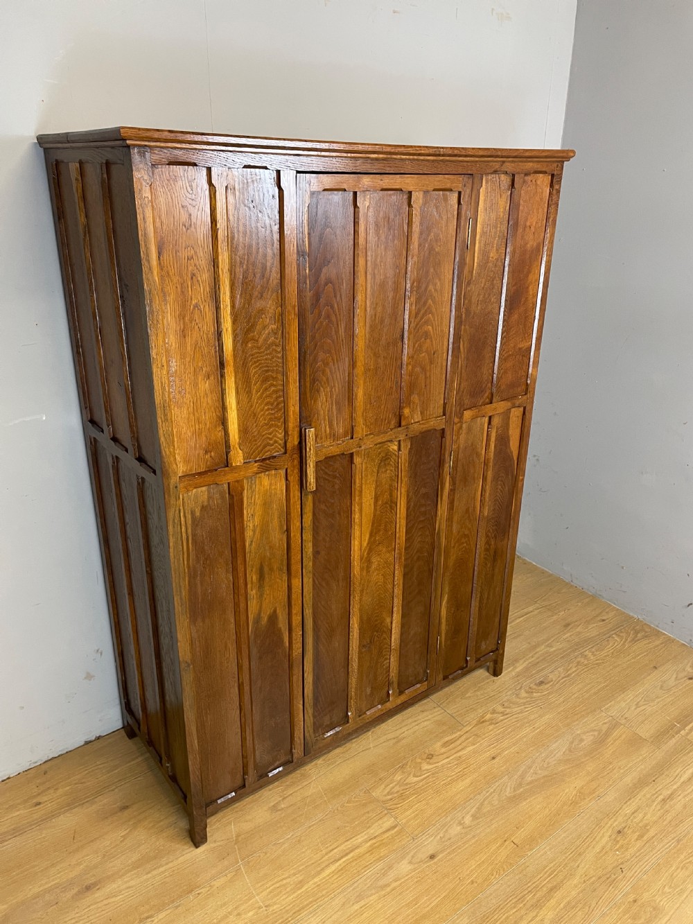 A solid warm brown oak wardrobe featuring vertical panelled double doors and exposed wooden handles in the Cotswold School style.