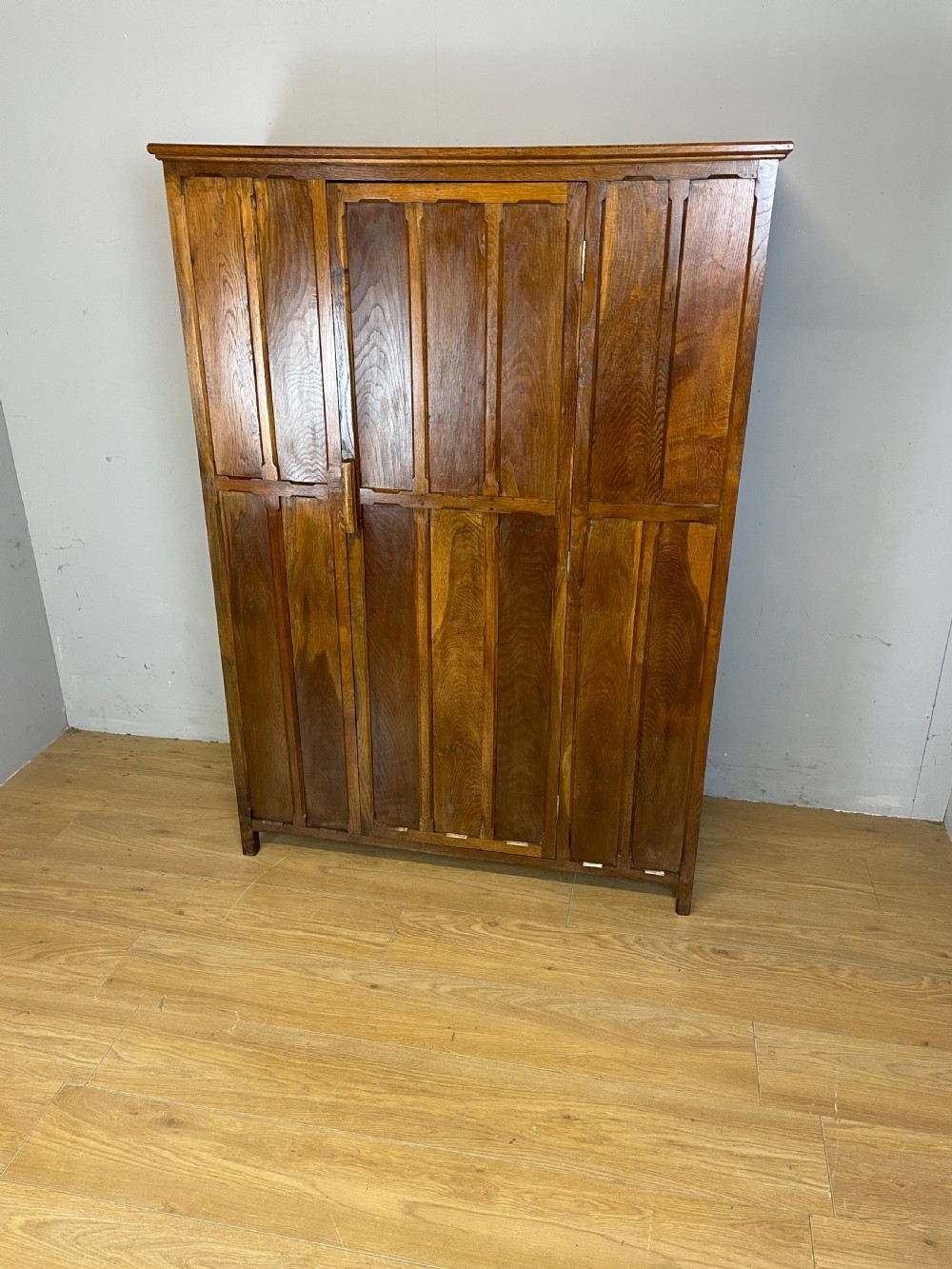 A solid warm brown oak wardrobe featuring vertical panelled double doors and exposed wooden handles in the Cotswold School style. View II