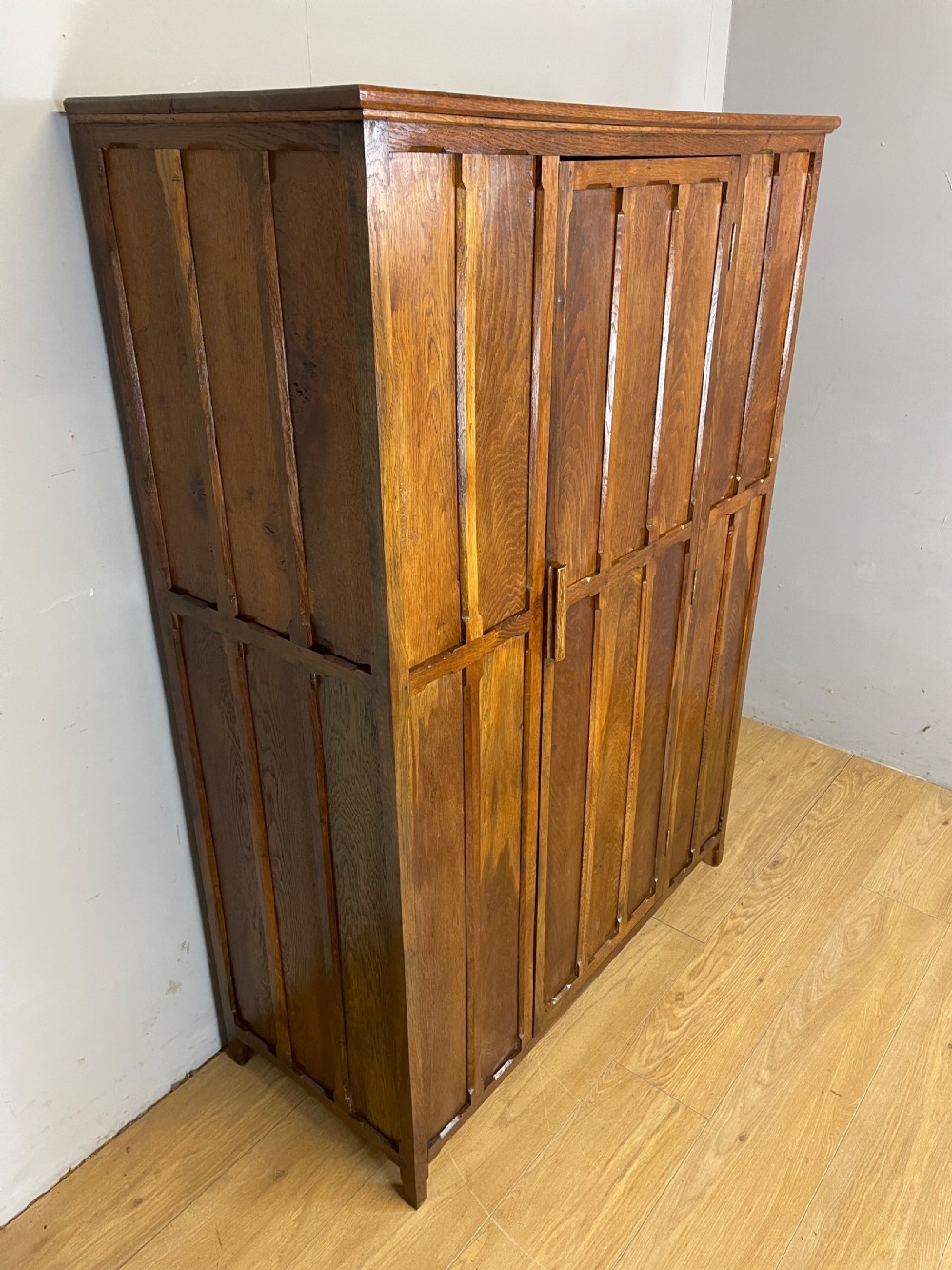 A solid warm brown oak wardrobe featuring vertical panelled double doors and exposed wooden handles in the Cotswold School style. View III