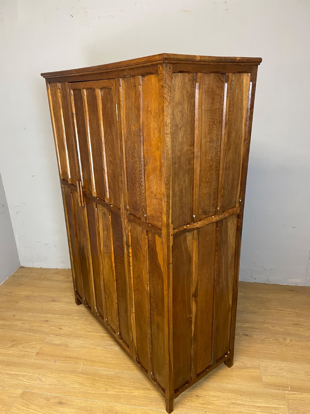 A solid warm brown oak wardrobe featuring vertical panelled double doors and exposed wooden handles in the Cotswold School style. View IX