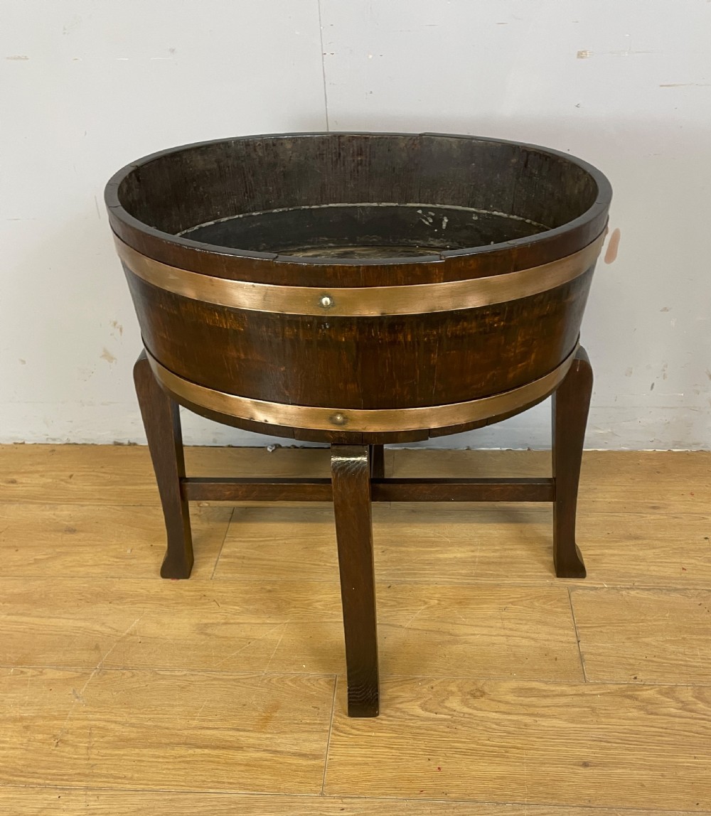 An Edwardian dark oak coopered circular planter with polished brass binding bands, raised on a three-legged wooden stand.