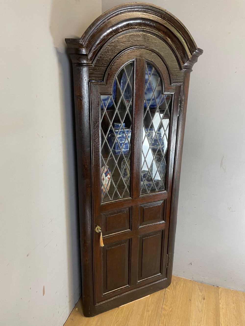 An early eighteenth-century dark oak corner cupboard featuring an arched top, leaded diamond glass door, and raised lower panels.