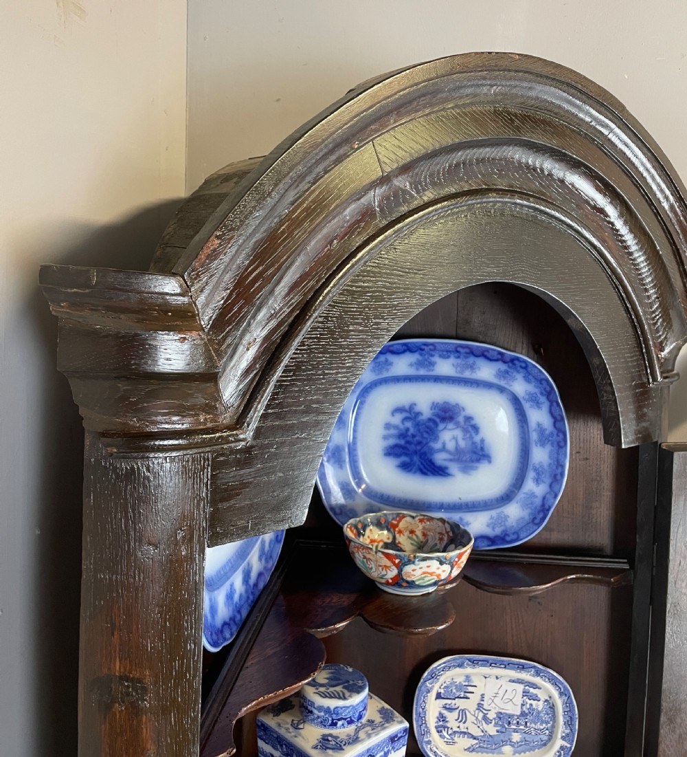 An early eighteenth-century dark oak corner cupboard featuring an arched top, leaded diamond glass door, and raised lower panels. View VI