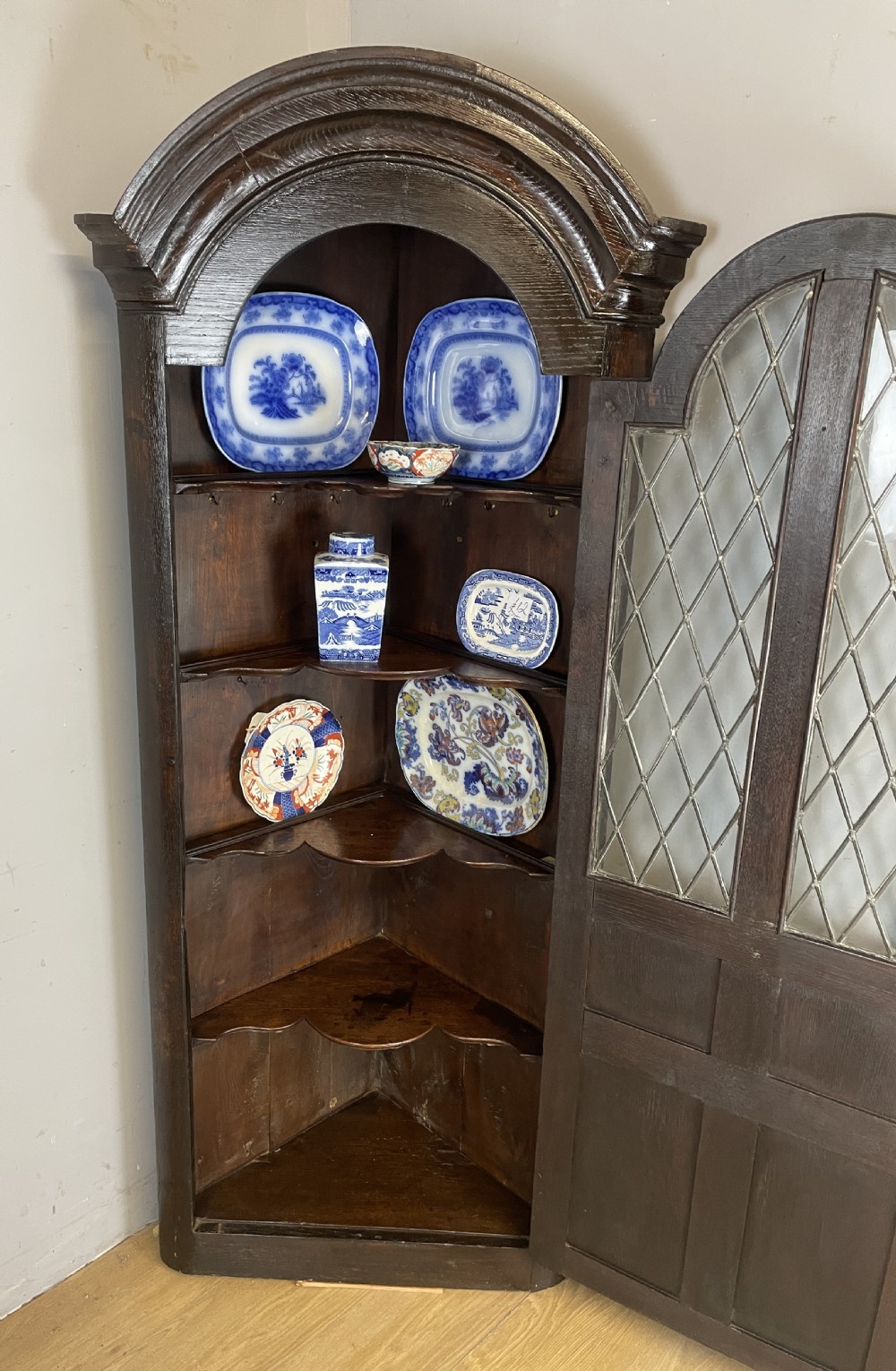 An early eighteenth-century dark oak corner cupboard featuring an arched top, leaded diamond glass door, and raised lower panels. View VII