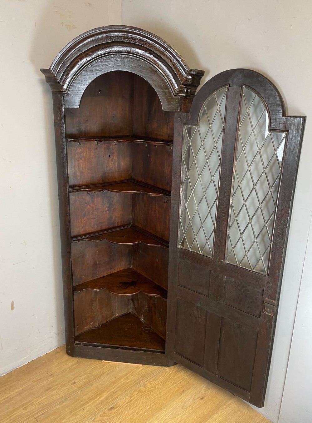 An early eighteenth-century dark oak corner cupboard featuring an arched top, leaded diamond glass door, and raised lower panels. View IX