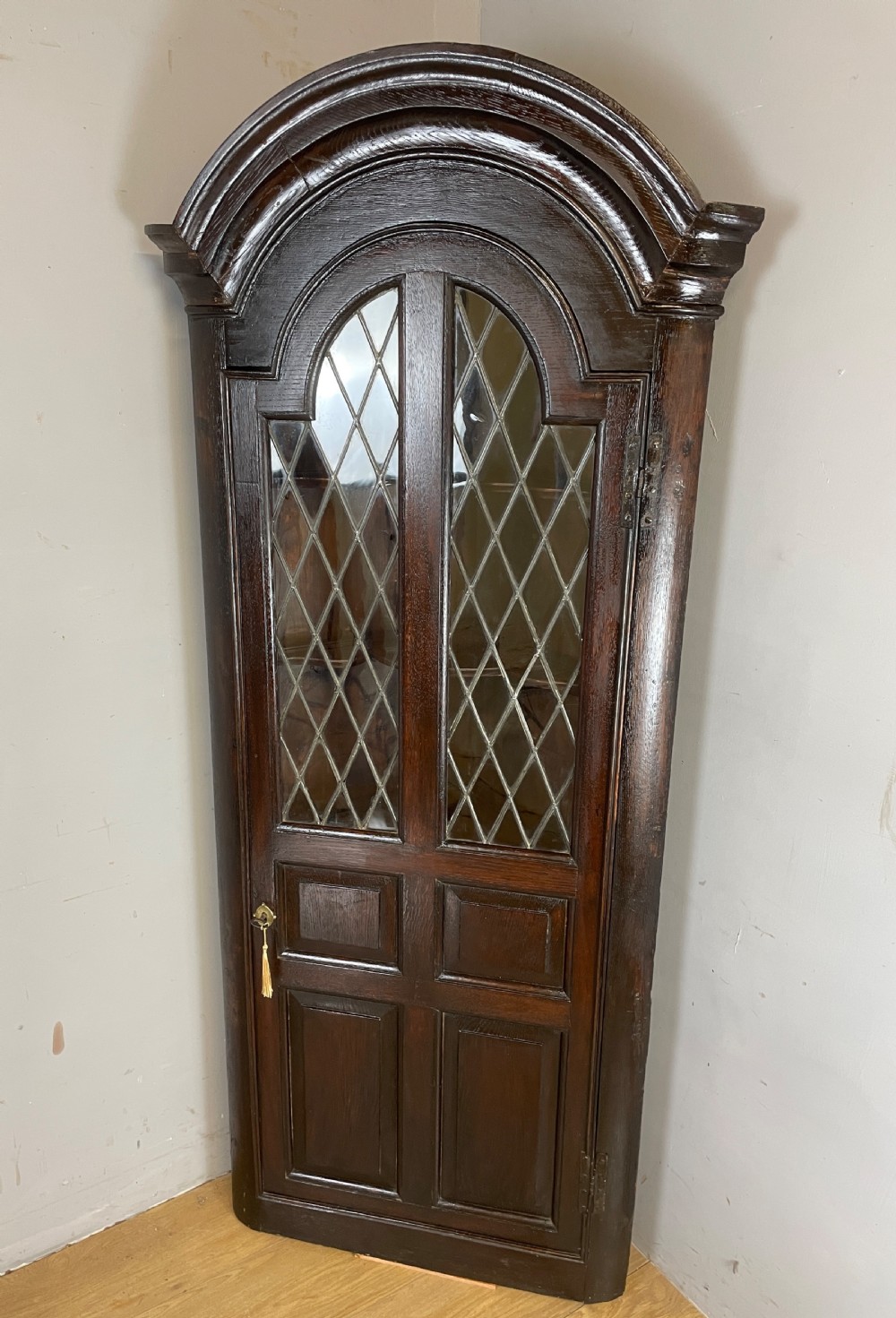 An early eighteenth-century dark oak corner cupboard featuring an arched top, leaded diamond glass door, and raised lower panels. View XI