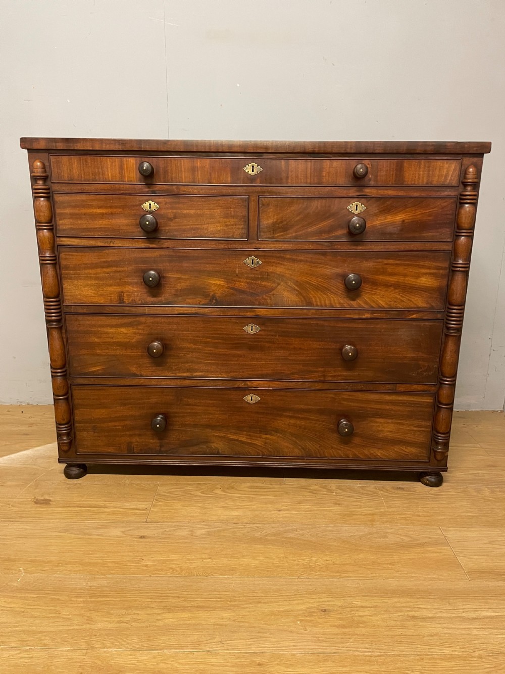 A Late Regency flame mahogany five-height chest of drawers with turned side columns, turned wooden knobs, and brass escutcheons. View VI
