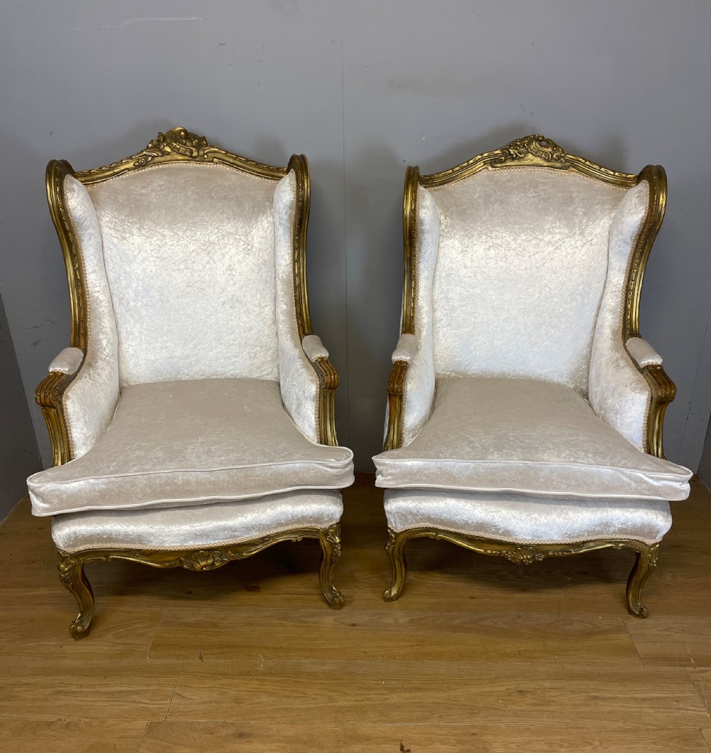 A pair of early twentieth-century French winged armchairs with carved gilded wooden frames and cream velvet upholstery.