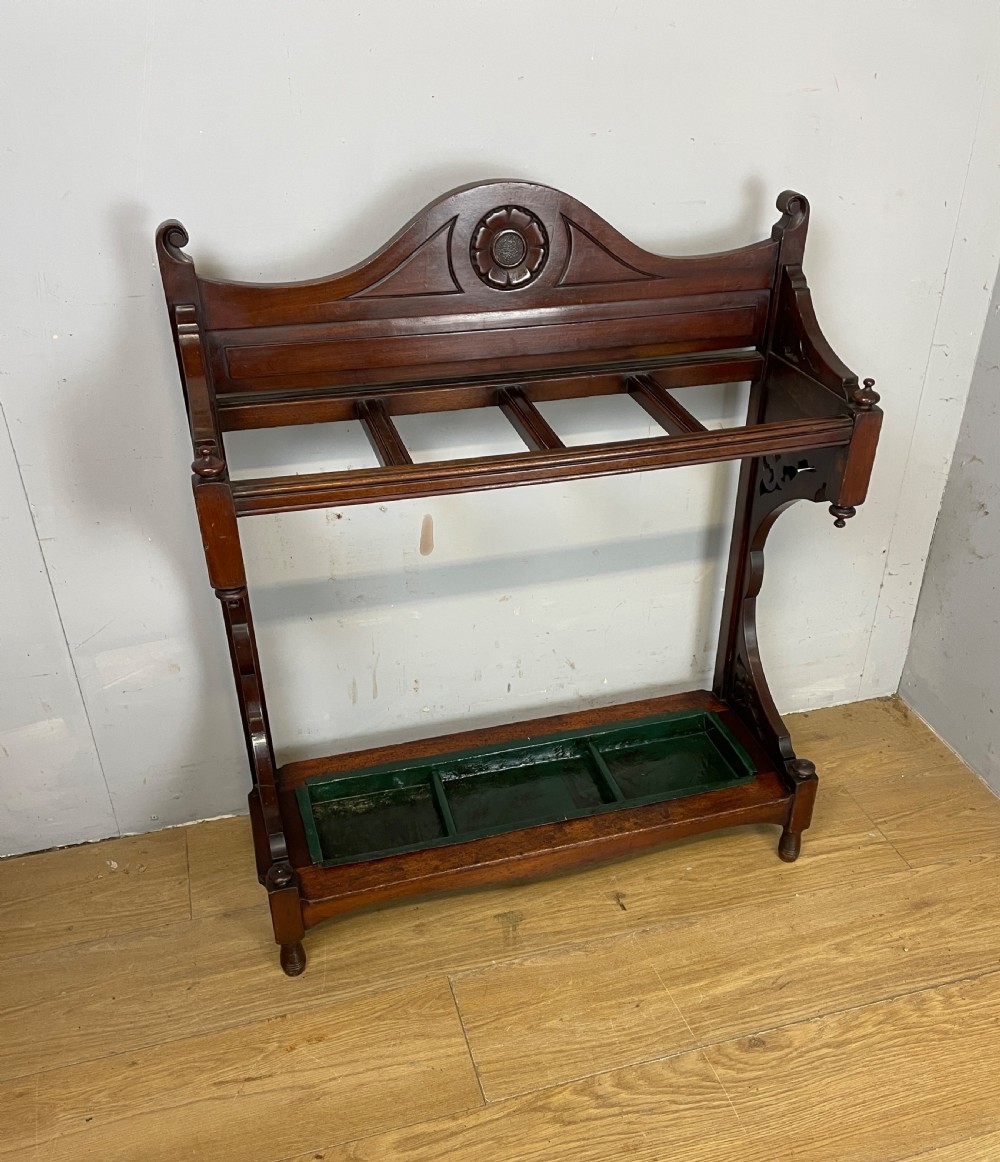 An Arts and Crafts mahogany stick stand featuring a carved central floral motif, divided top rail, turned finials, and a removable green metal drip tray at the base.