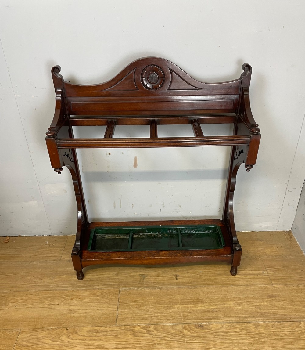 An Arts and Crafts mahogany stick stand featuring a carved central floral motif, divided top rail, turned finials, and a removable green metal drip tray at the base. View II