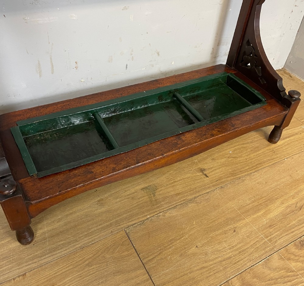 An Arts and Crafts mahogany stick stand featuring a carved central floral motif, divided top rail, turned finials, and a removable green metal drip tray at the base. View III