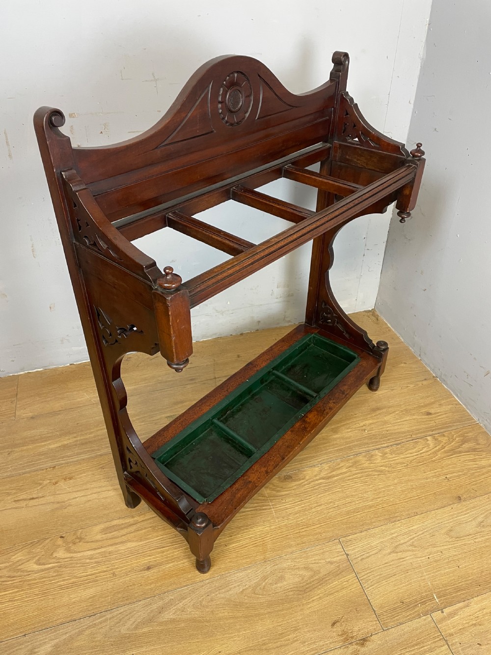 An Arts and Crafts mahogany stick stand featuring a carved central floral motif, divided top rail, turned finials, and a removable green metal drip tray at the base. View IV
