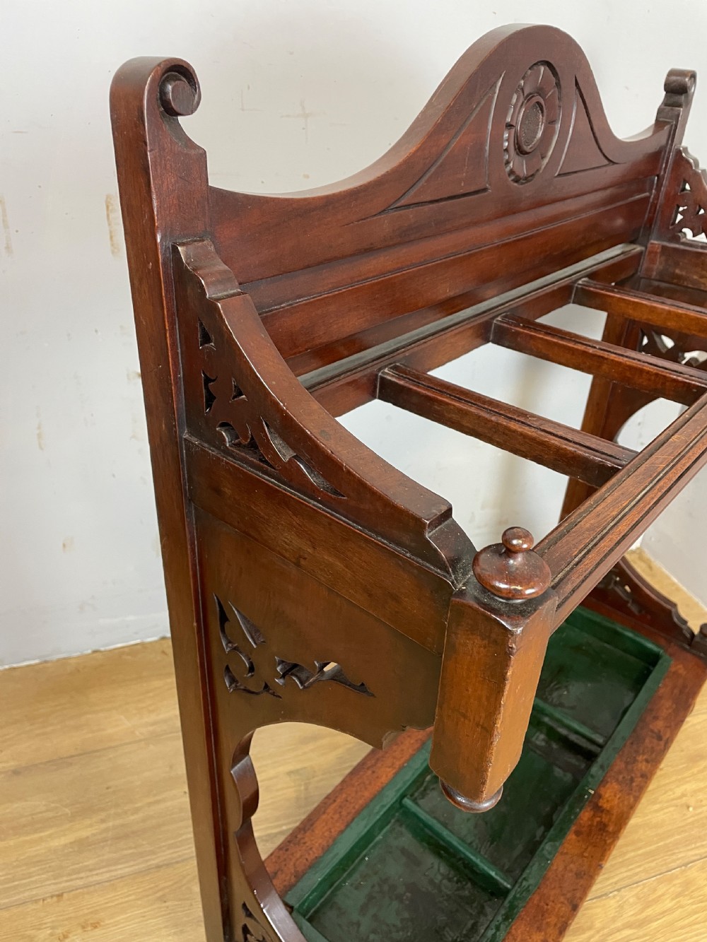 An Arts and Crafts mahogany stick stand featuring a carved central floral motif, divided top rail, turned finials, and a removable green metal drip tray at the base. View V