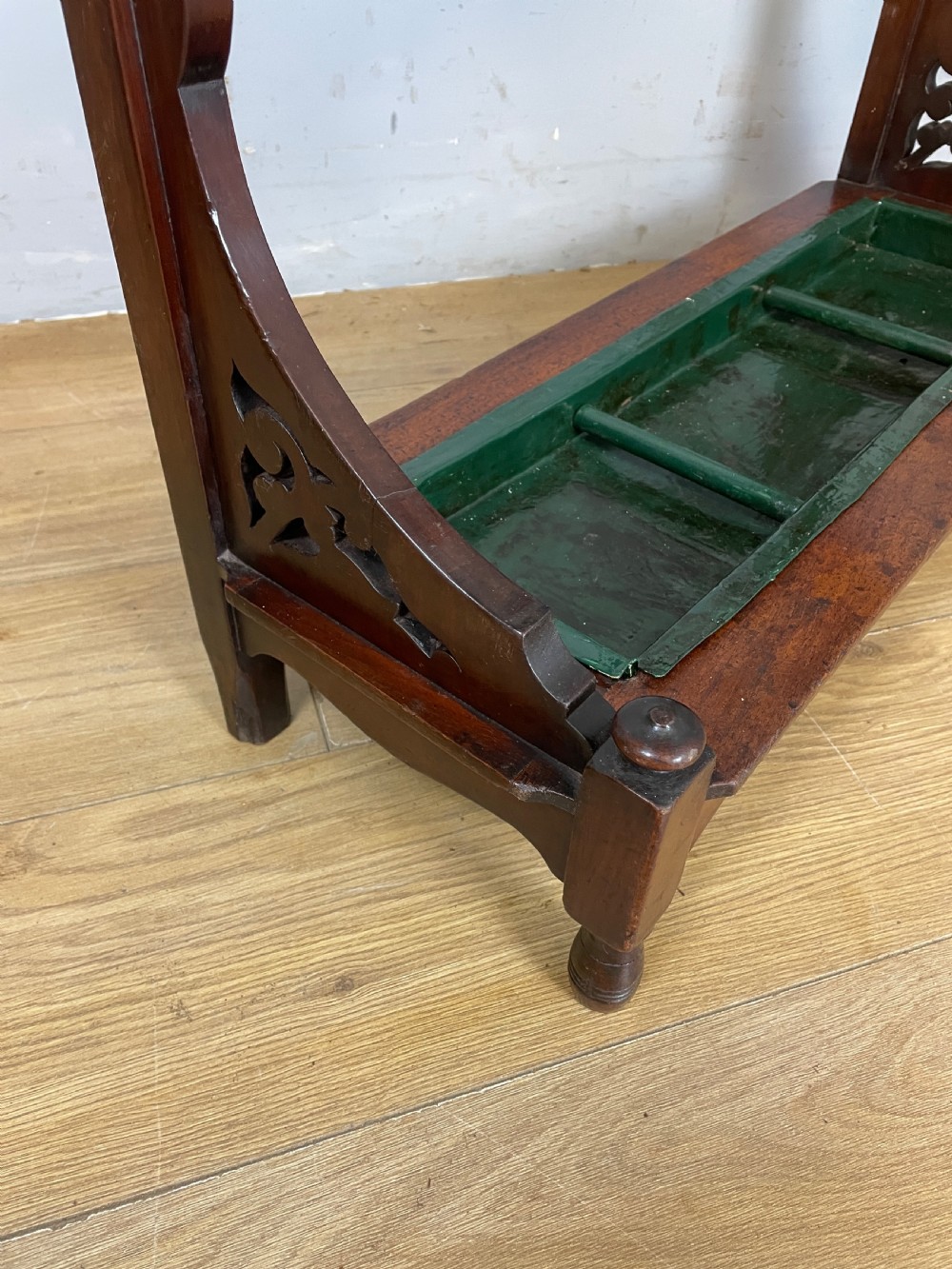 An Arts and Crafts mahogany stick stand featuring a carved central floral motif, divided top rail, turned finials, and a removable green metal drip tray at the base. View VI