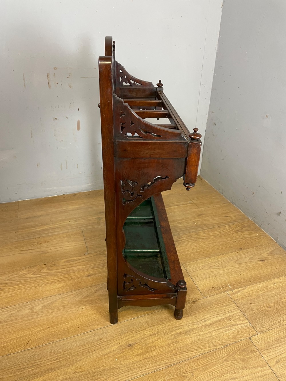 An Arts and Crafts mahogany stick stand featuring a carved central floral motif, divided top rail, turned finials, and a removable green metal drip tray at the base. View VII
